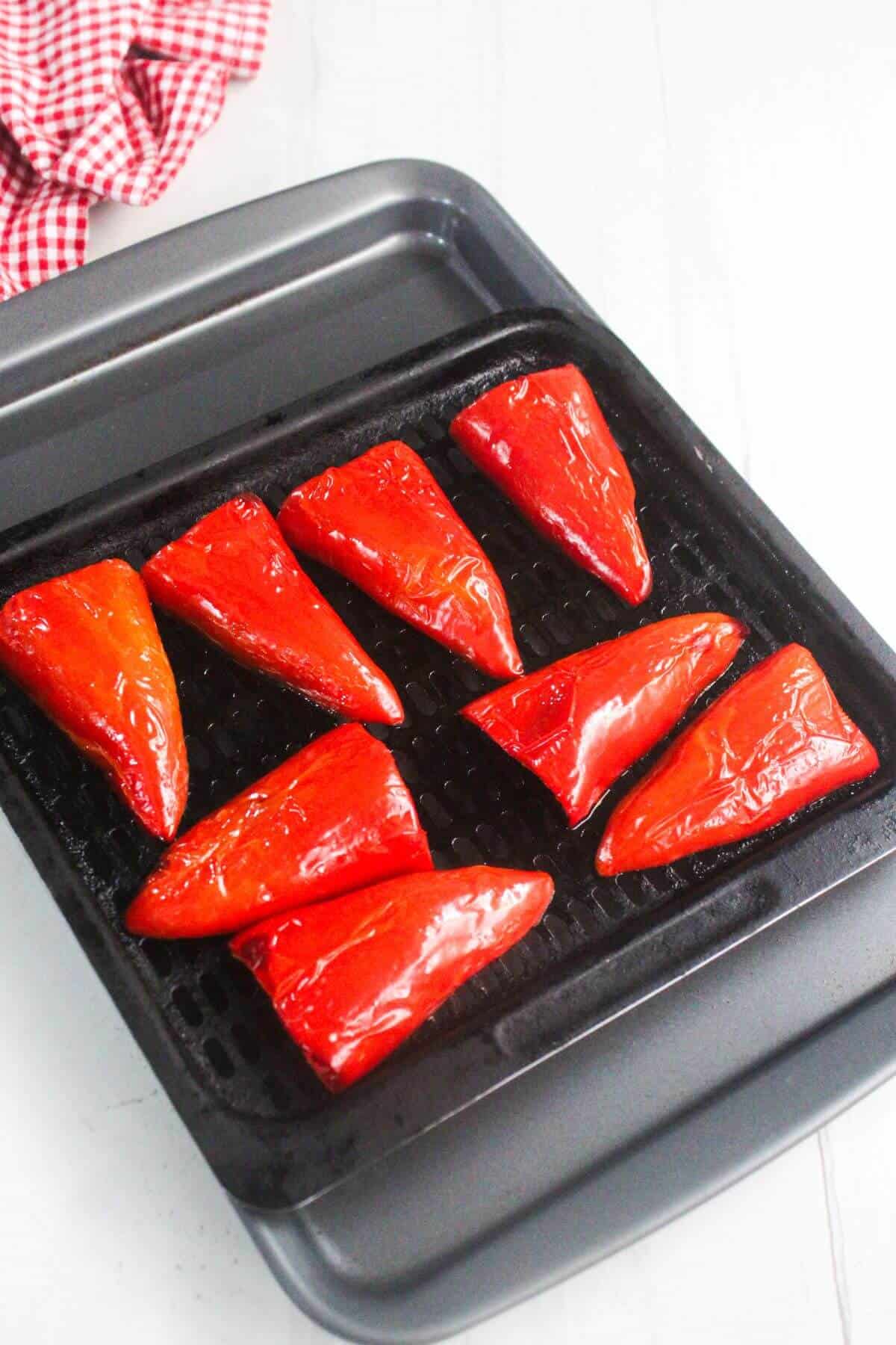 Red bell pepper halves arranged on a black grill pan, ready to be roasted, with a red checkered cloth in the corner.
