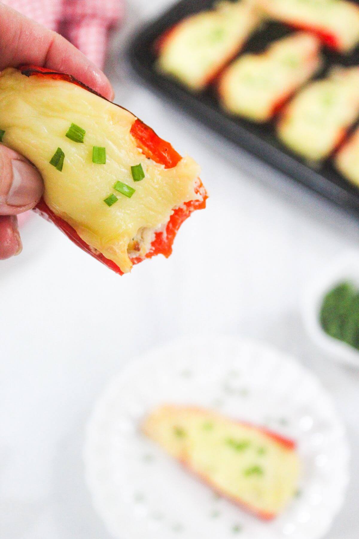 A hand holds a stuffed red bell pepper topped with melted cheese and chopped chives; more stuffed peppers are on a tray in the background.