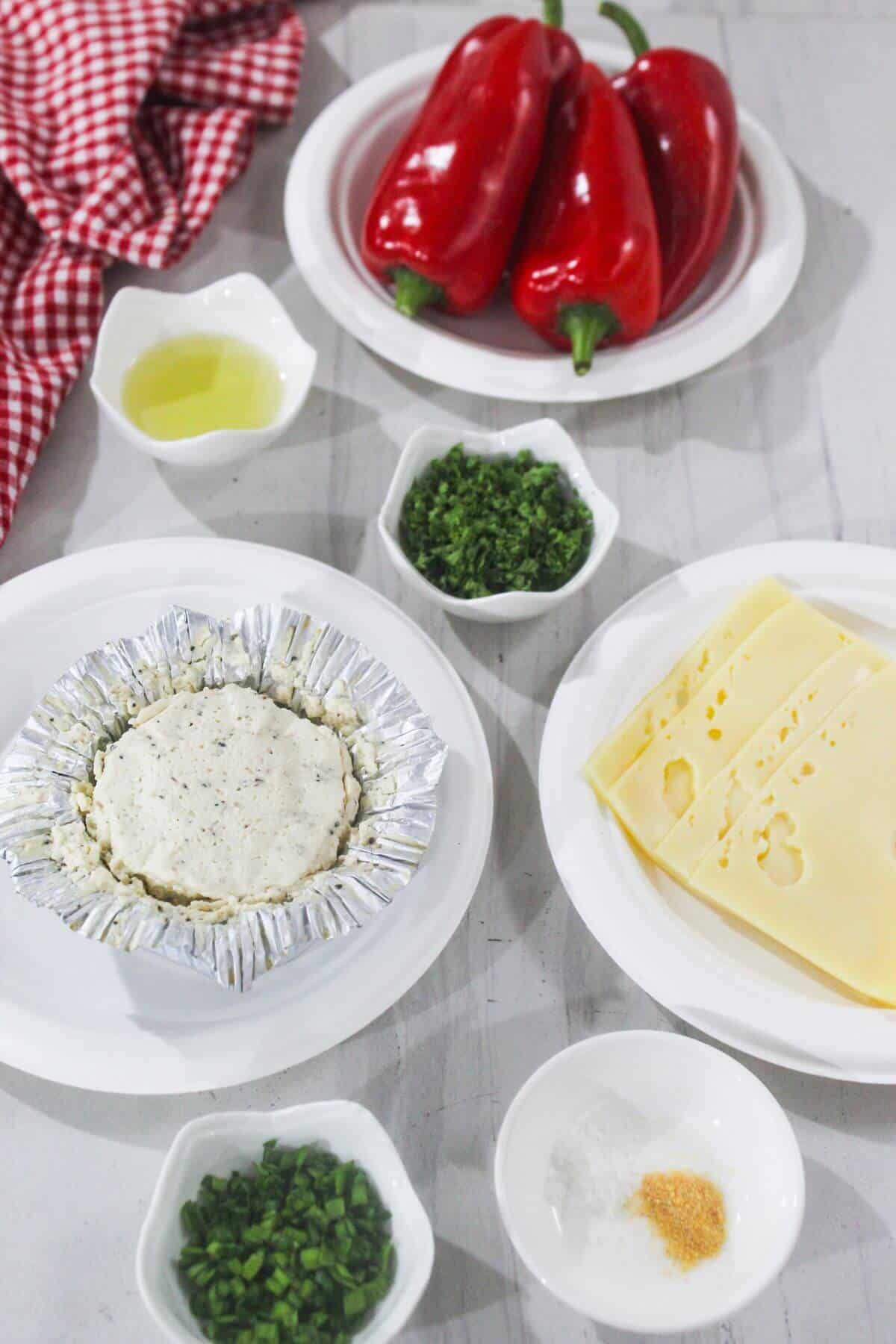 Several plates and bowls hold ingredients including red bell peppers, sliced cheese, herbs, oil, spices, and a round foil-wrapped cheese on a white surface with a red checkered cloth.