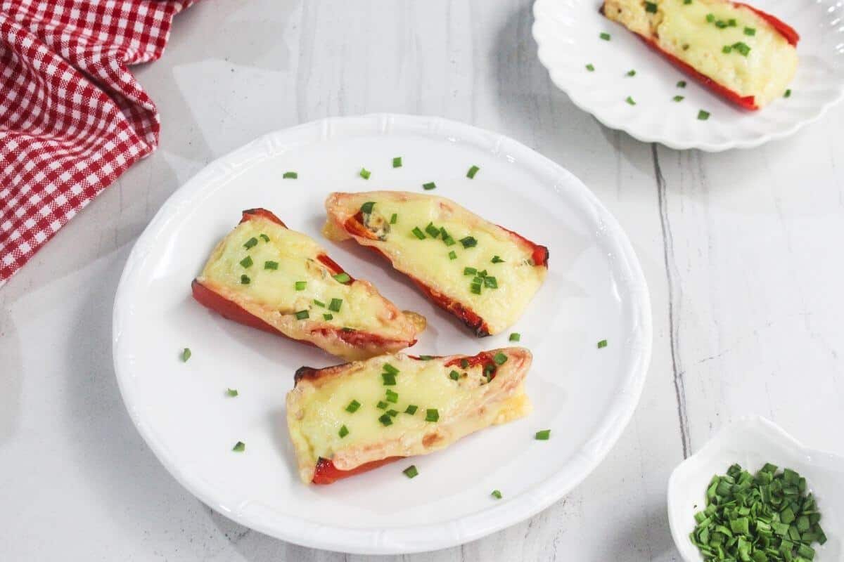 Three stuffed red bell pepper halves topped with melted cheese and chopped chives on a white plate, with a small bowl of chives and a red checkered cloth nearby.