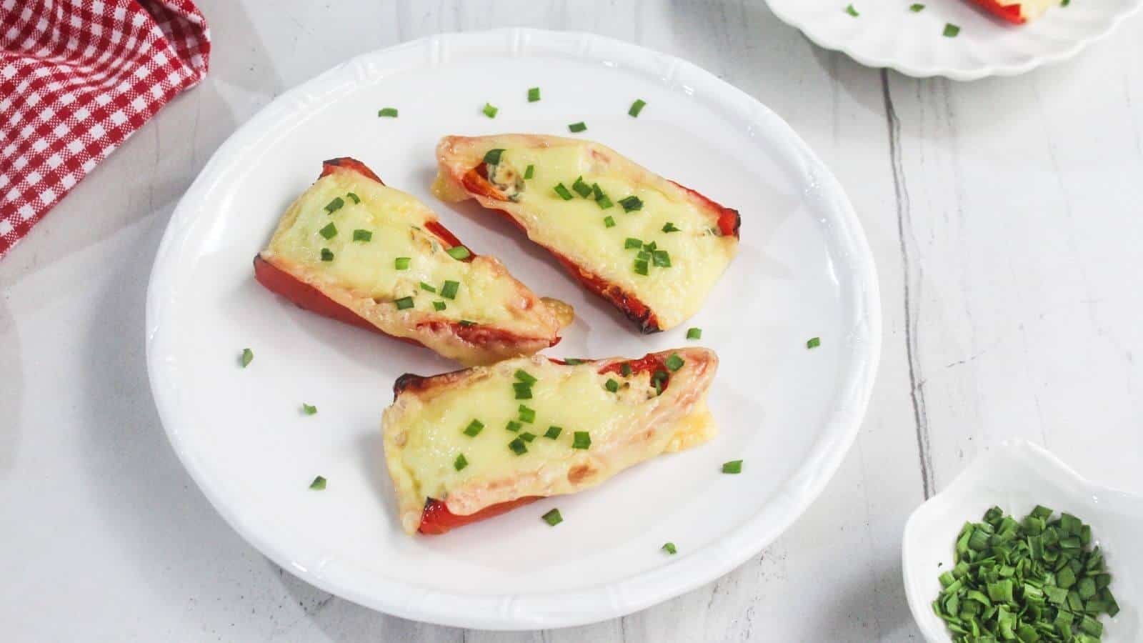 Three stuffed red bell pepper halves topped with melted cheese and chopped chives on a white plate, with a small bowl of chives nearby.