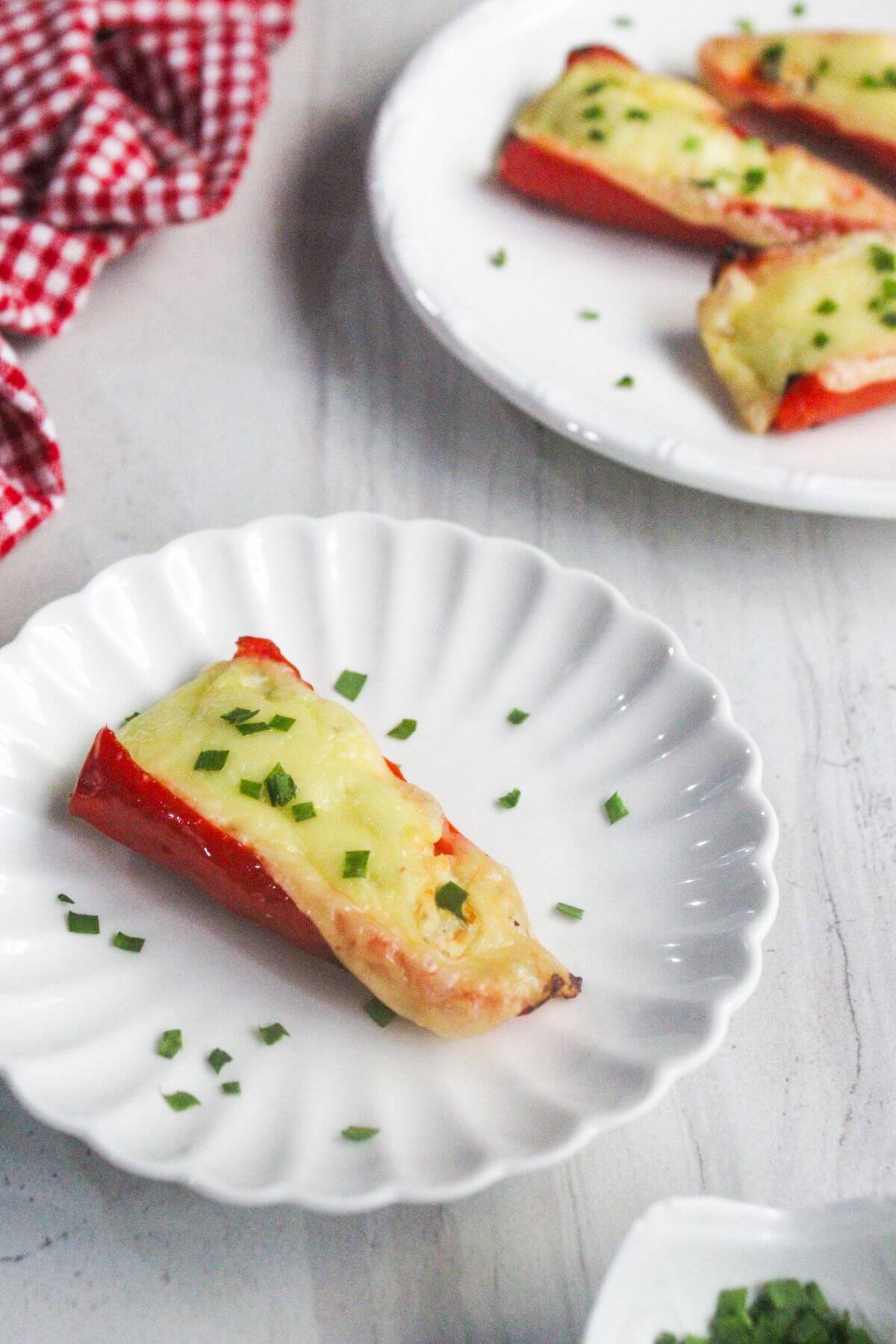 A red bell pepper half stuffed with melted cheese and garnished with chopped chives on a white plate, with more stuffed peppers in the background.