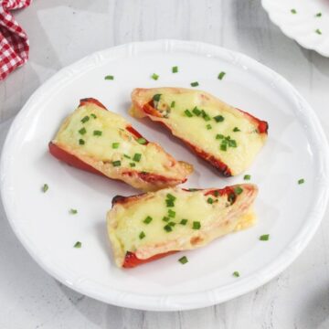 Three stuffed red bell pepper halves topped with melted cheese and chopped green herbs are arranged on a white plate.
