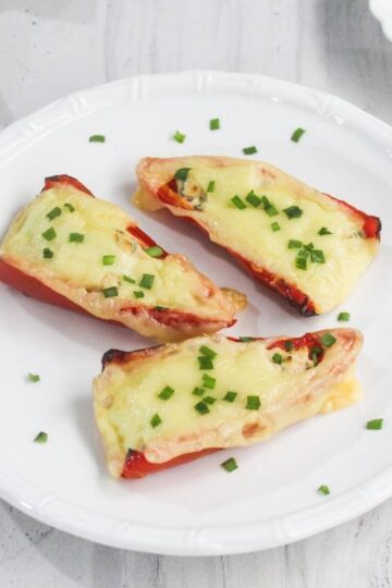 Three stuffed red bell pepper halves topped with melted cheese and chopped green herbs are arranged on a white plate.