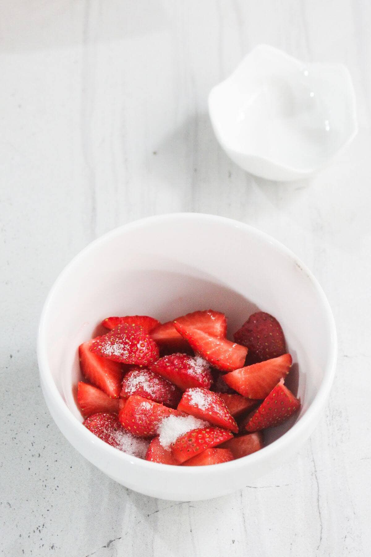 A white bowl filled with sliced strawberries sprinkled with sugar sits on a light marble surface; a small empty white bowl is in the background.