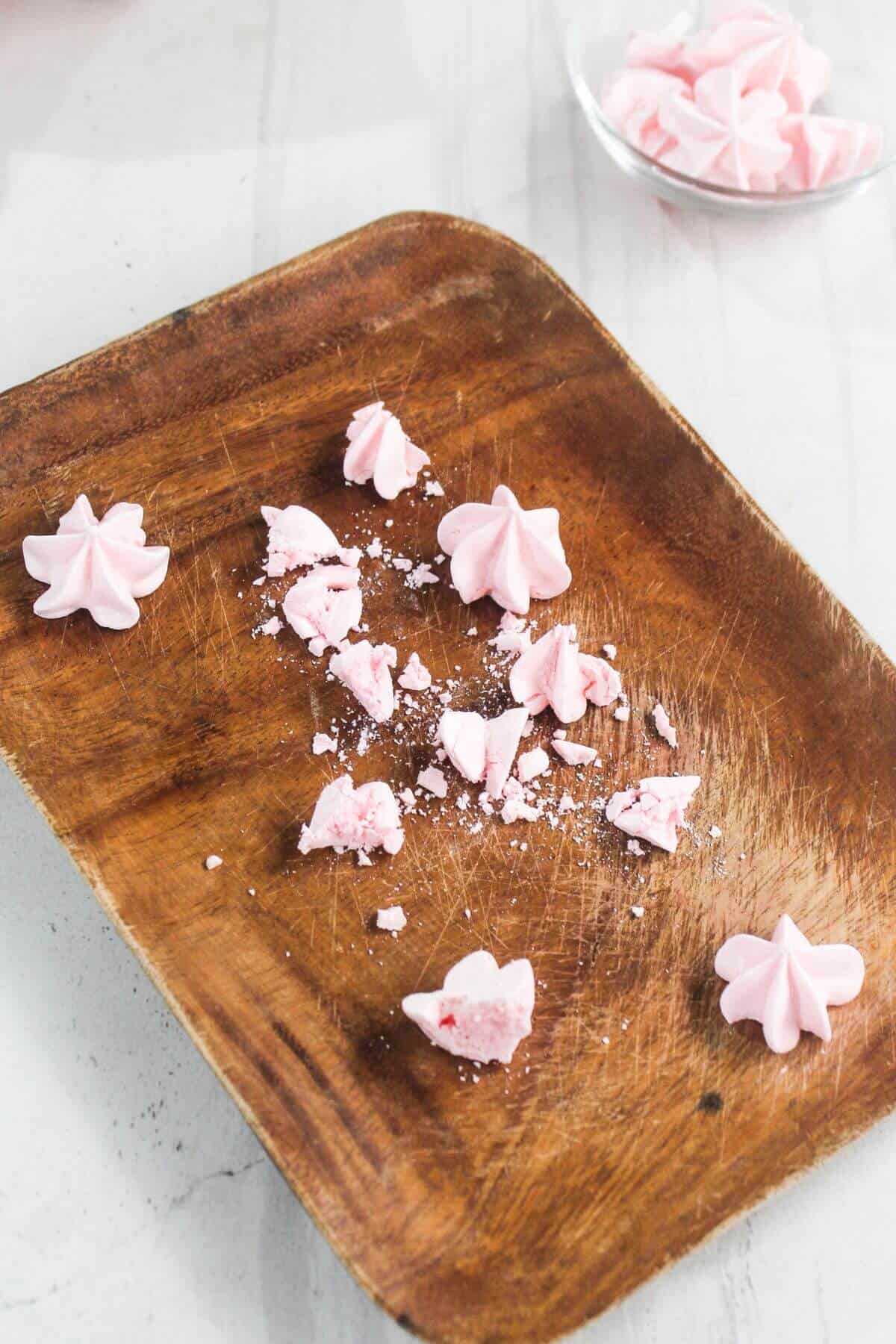 A wooden tray with several pink meringue cookies, some whole and some broken, on a light surface; a glass bowl with more meringues is in the background.