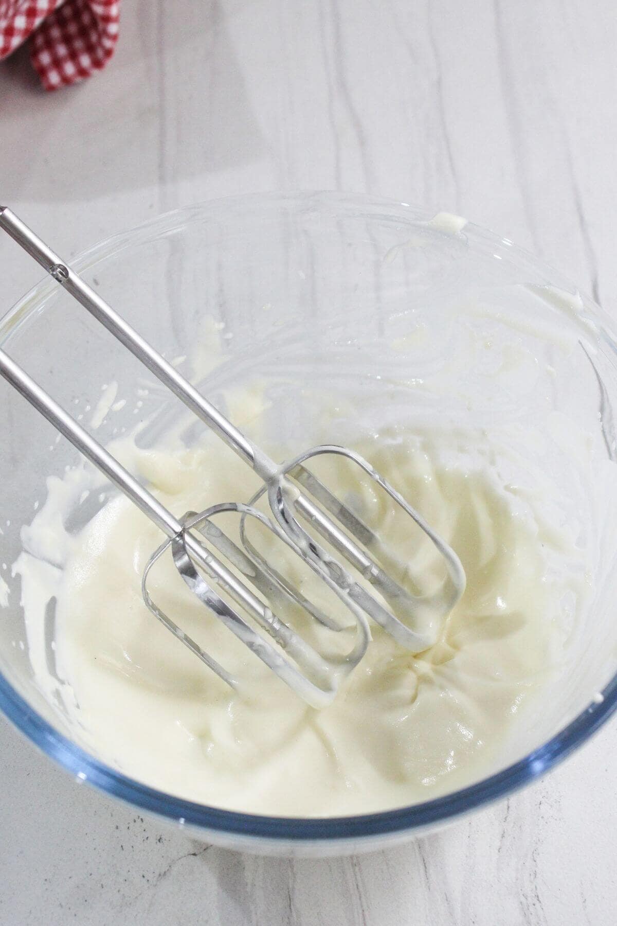 A glass bowl containing whipped cream and two metal beaters resting inside, on a white countertop.