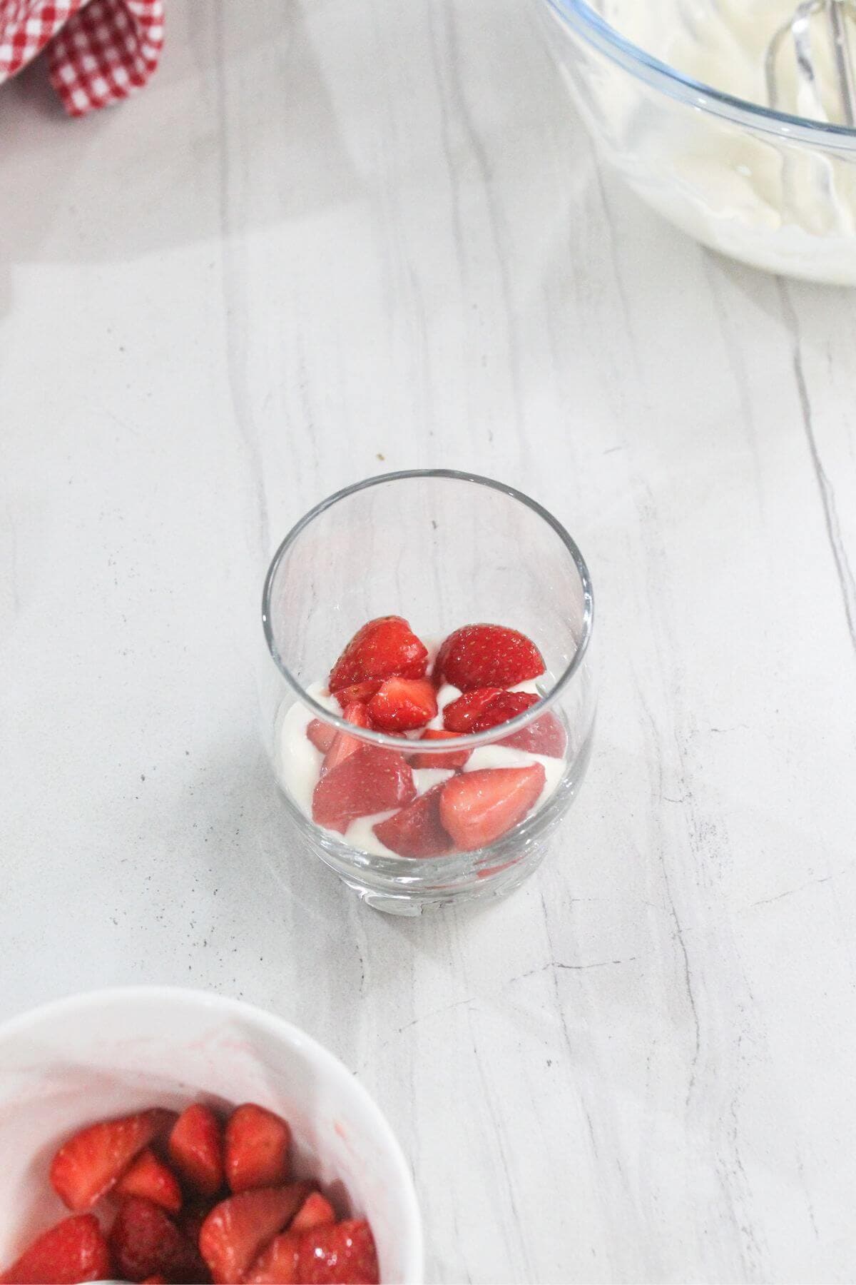 A clear glass with layers of sliced strawberries and cream sits on a white marble surface, with a bowl of strawberries and a glass bowl of cream nearby.