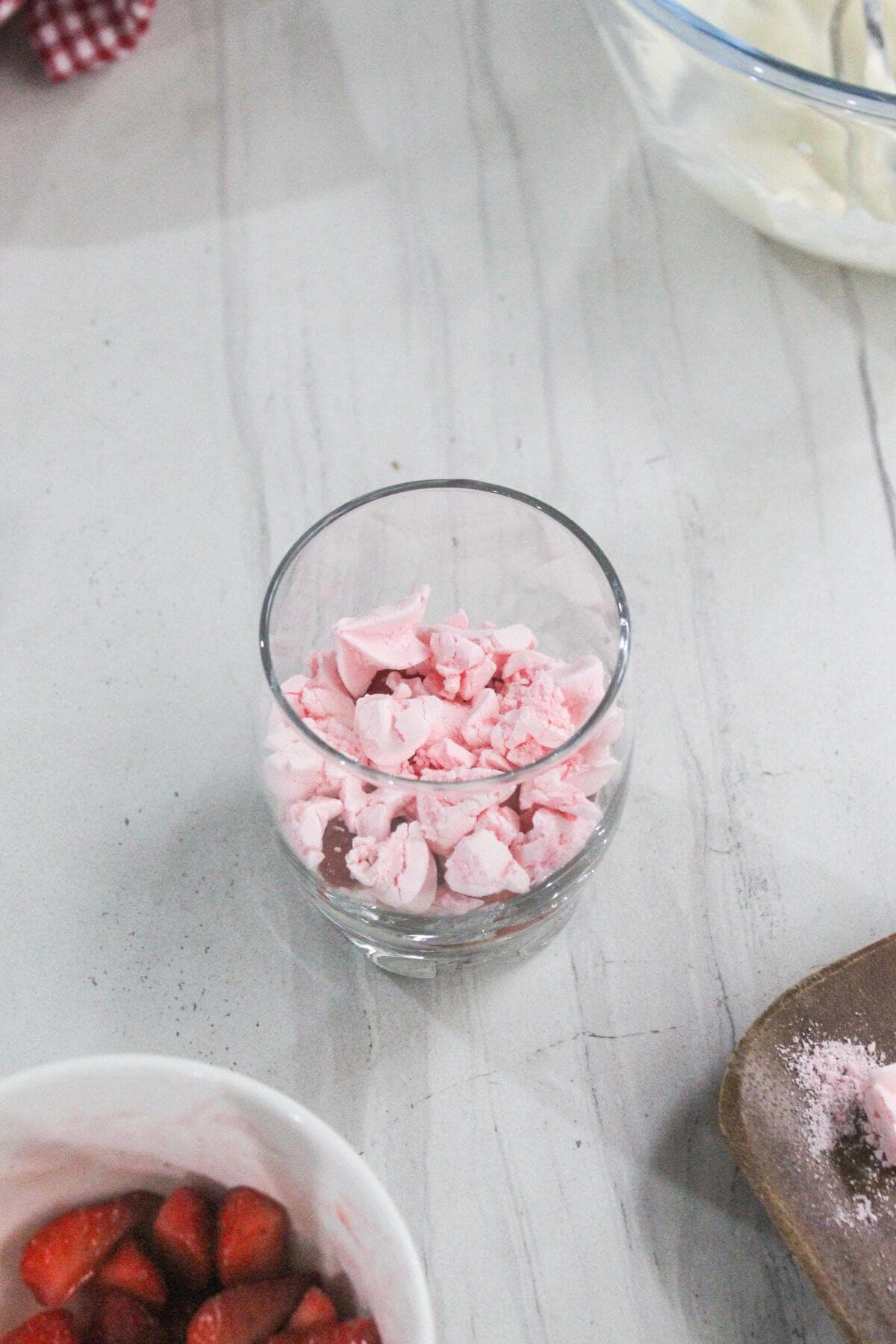 A glass filled with pink meringue pieces sits on a light-colored countertop next to a bowl of sliced strawberries and a partially visible plate.