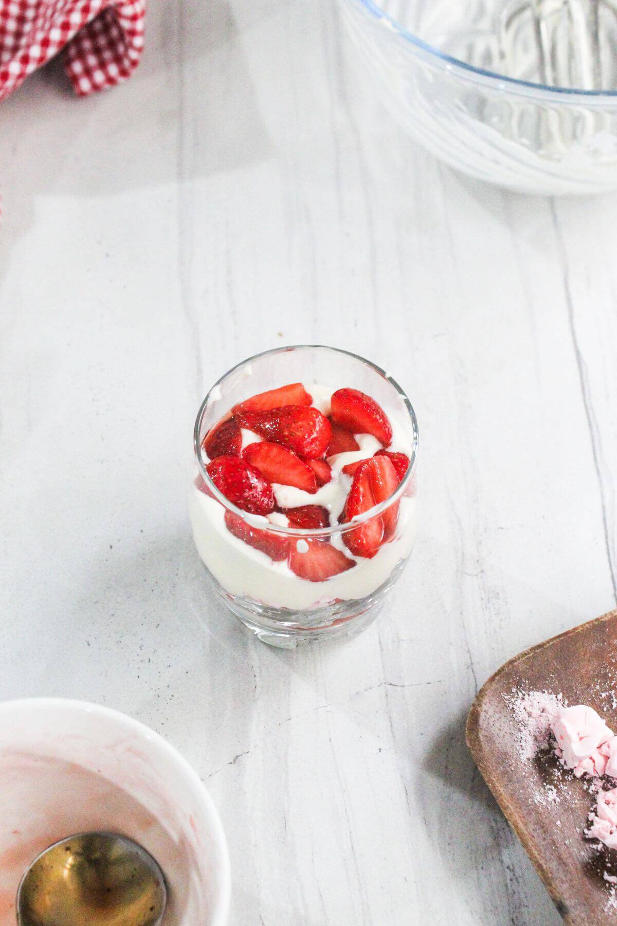 A glass filled with layers of white yogurt and sliced strawberries sits on a white surface near an empty bowl and a dirty spoon.