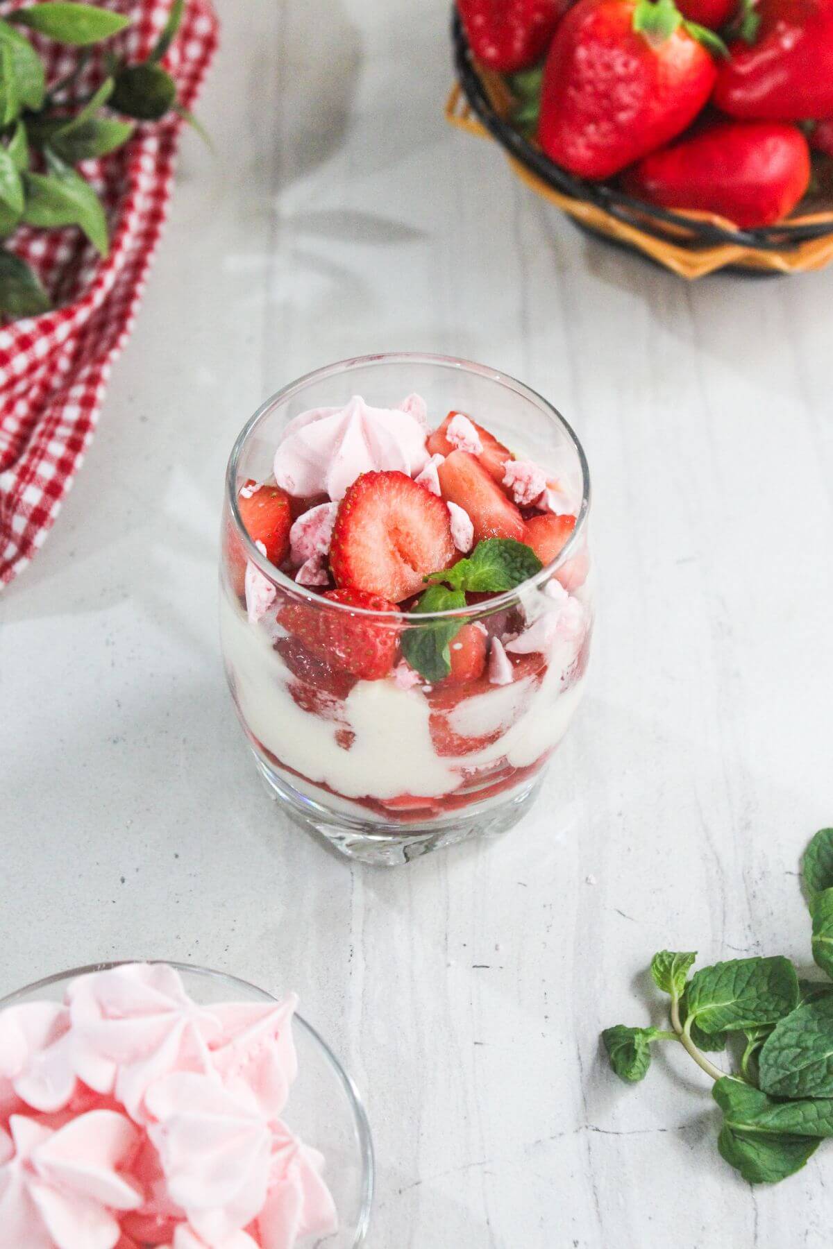 A glass of layered dessert with cream, sliced strawberries, and pink meringue pieces, garnished with mint leaves. Fresh strawberries and mint are on the table nearby.