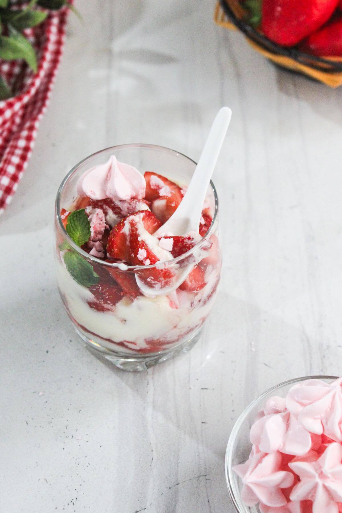 A glass filled with layers of whipped cream, strawberries, and pink meringue pieces, with a white spoon inside. A bowl of meringues and a basket of strawberries are nearby.