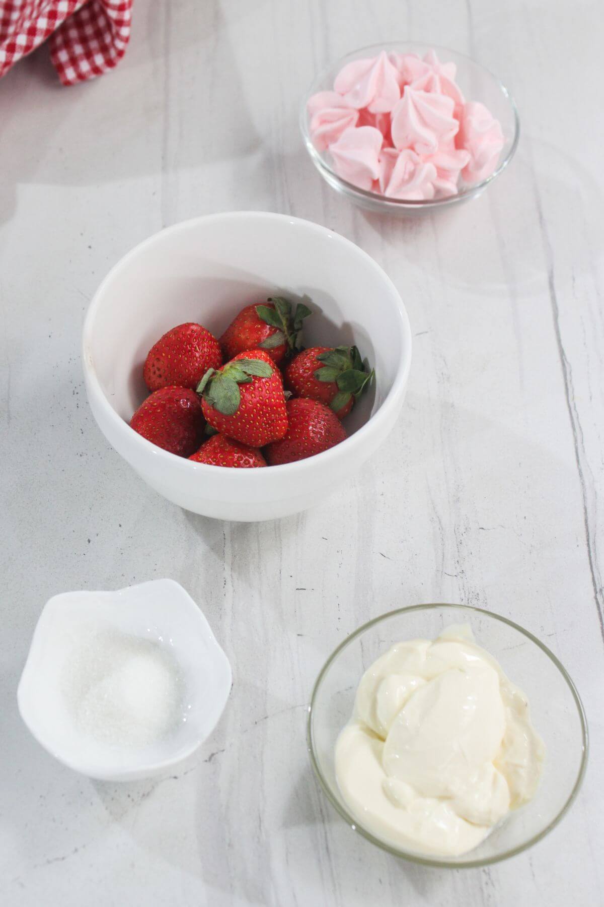 A white bowl of strawberries, a small bowl of sugar, a dish of whipped cream, and a bowl of pink meringues on a light surface.