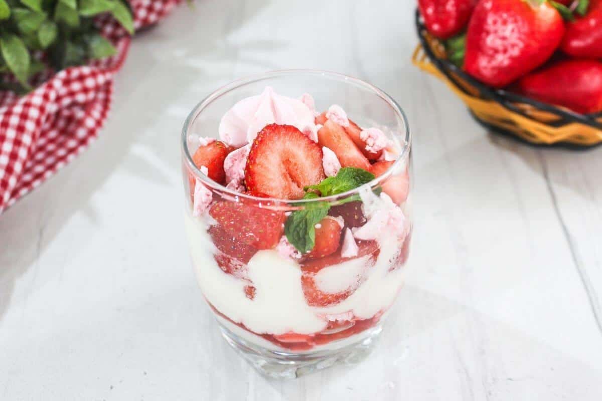 A glass filled with layers of sliced strawberries, whipped cream, and meringue, garnished with mint, sits on a white surface next to a basket of fresh strawberries.