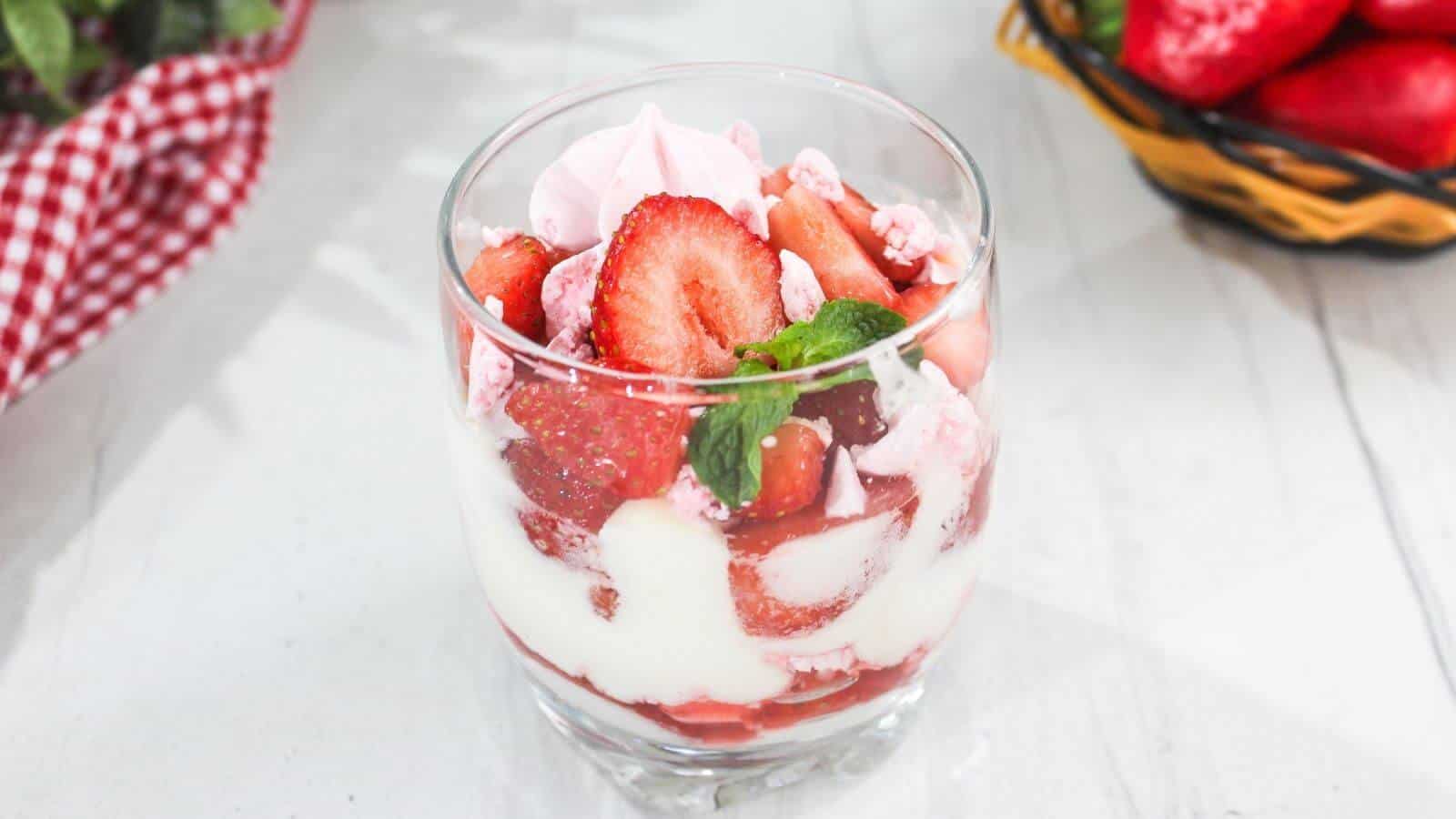 A glass filled with layers of strawberries, cream, and meringue pieces, garnished with mint, sits on a white surface next to baskets of strawberries.