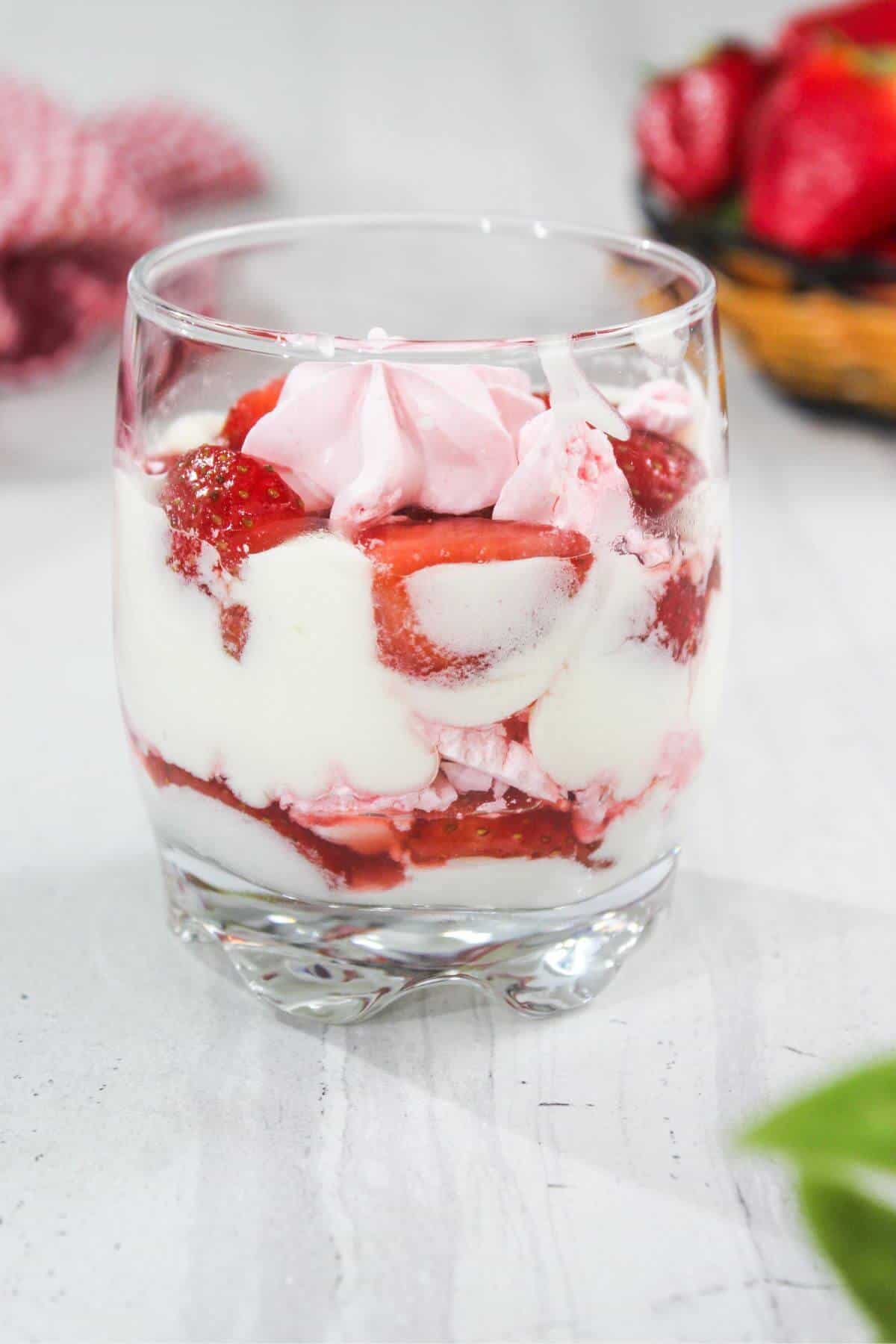 A glass filled with layers of whipped cream, strawberry sauce, fresh strawberries, and pink meringue on a light surface.