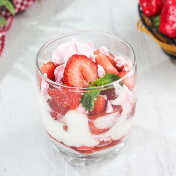 A glass filled with layers of sliced strawberries, white cream, and pink meringue pieces, garnished with a mint leaf, on a white surface.