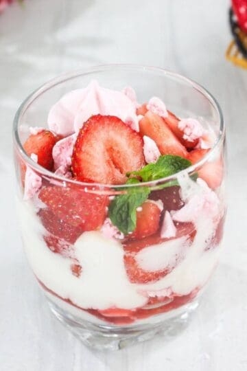 A glass filled with layers of sliced strawberries, white cream, and pink meringue pieces, garnished with a mint leaf, on a white surface.