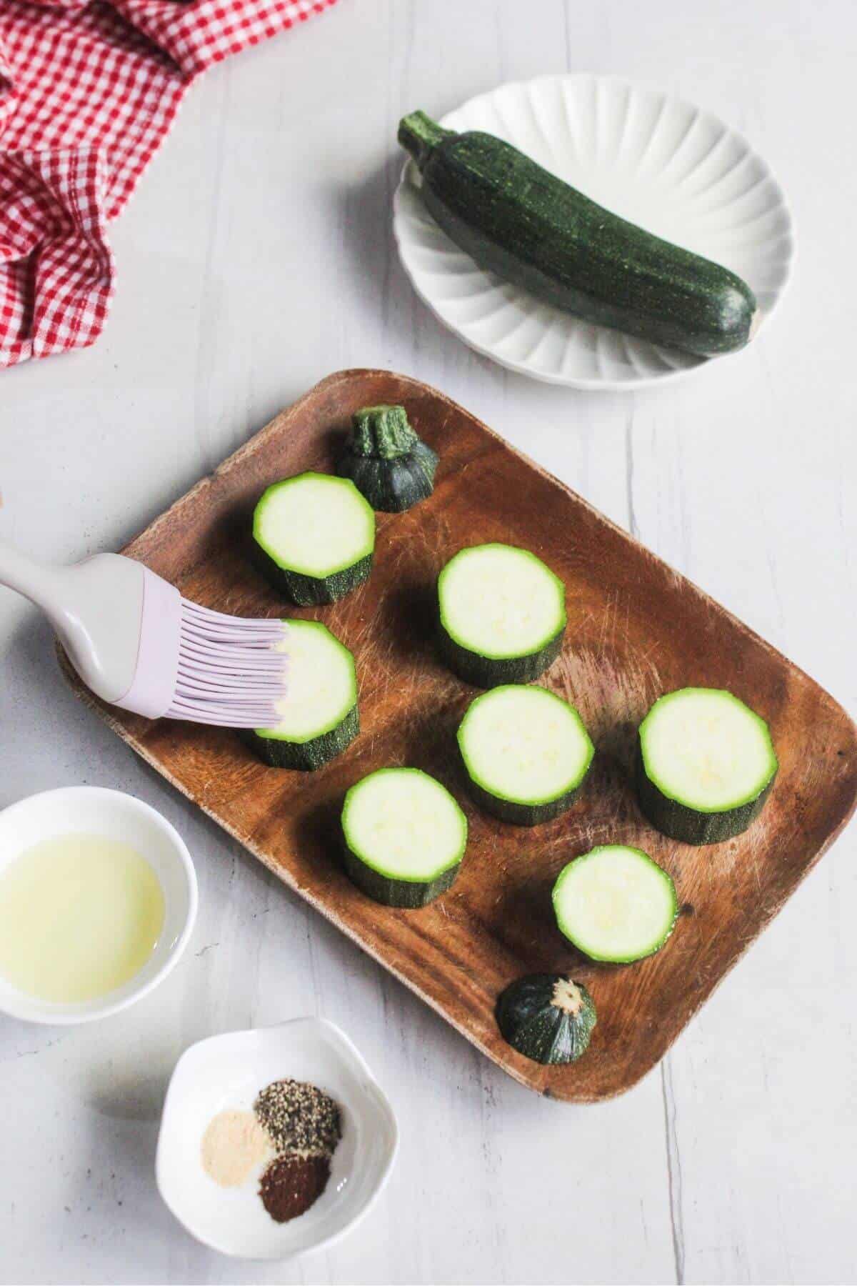 Sliced zucchini on a wooden tray being brushed with oil, with whole zucchini, spices, and oil in small bowls on a white table.
