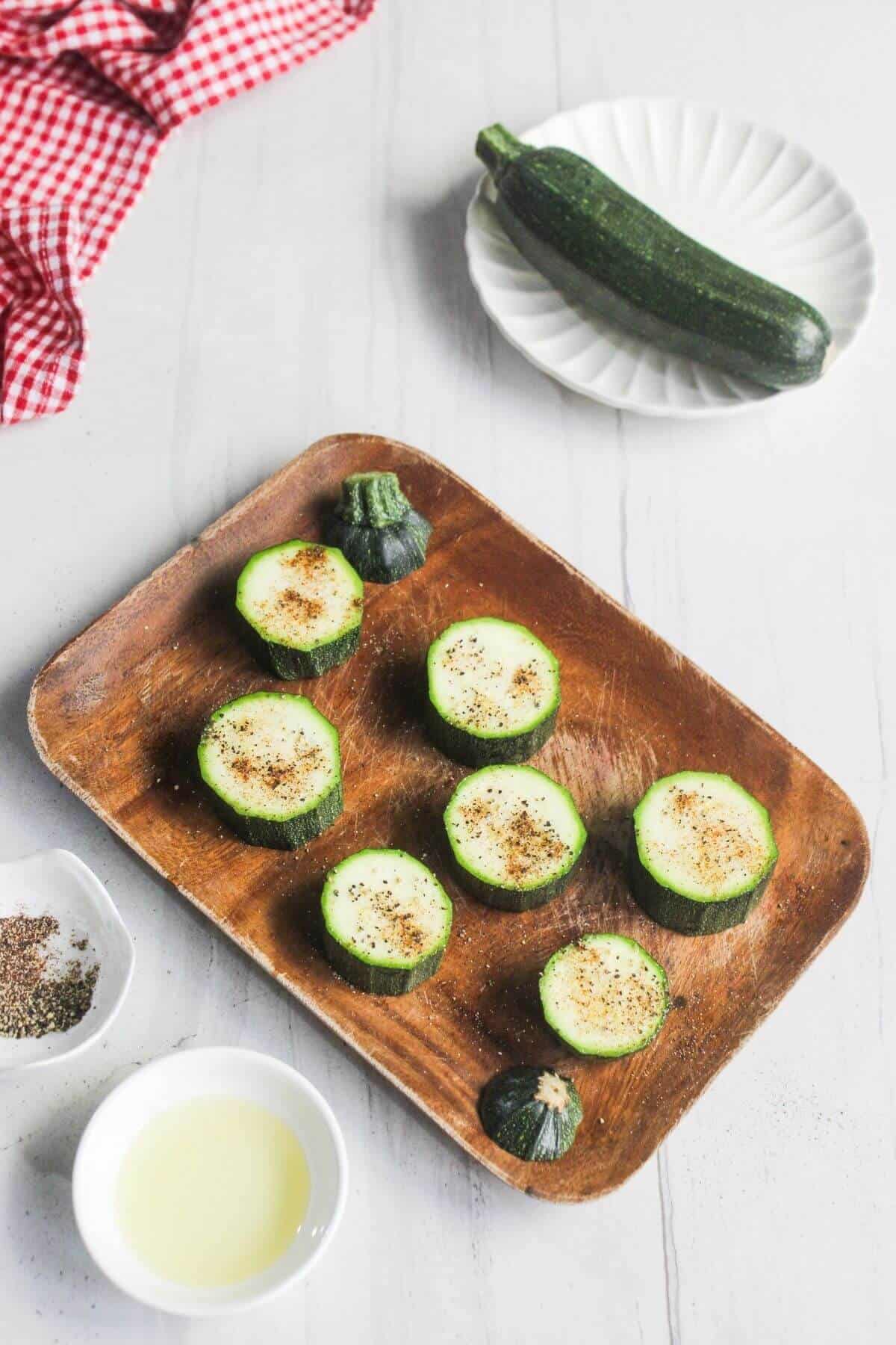 Sliced zucchini rounds on a wooden tray, seasoned with pepper, next to bowls of oil and pepper, with a whole zucchini on a plate and a red checkered cloth in the background.