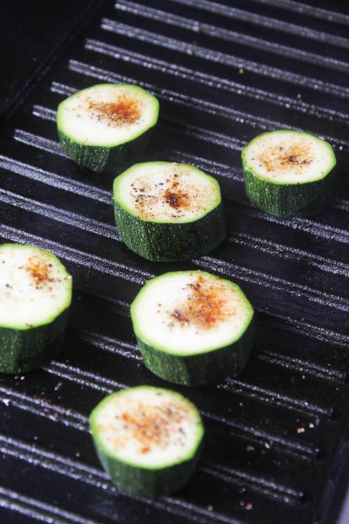 Sliced zucchini rounds seasoned with spices are cooking on a ridged grill pan.