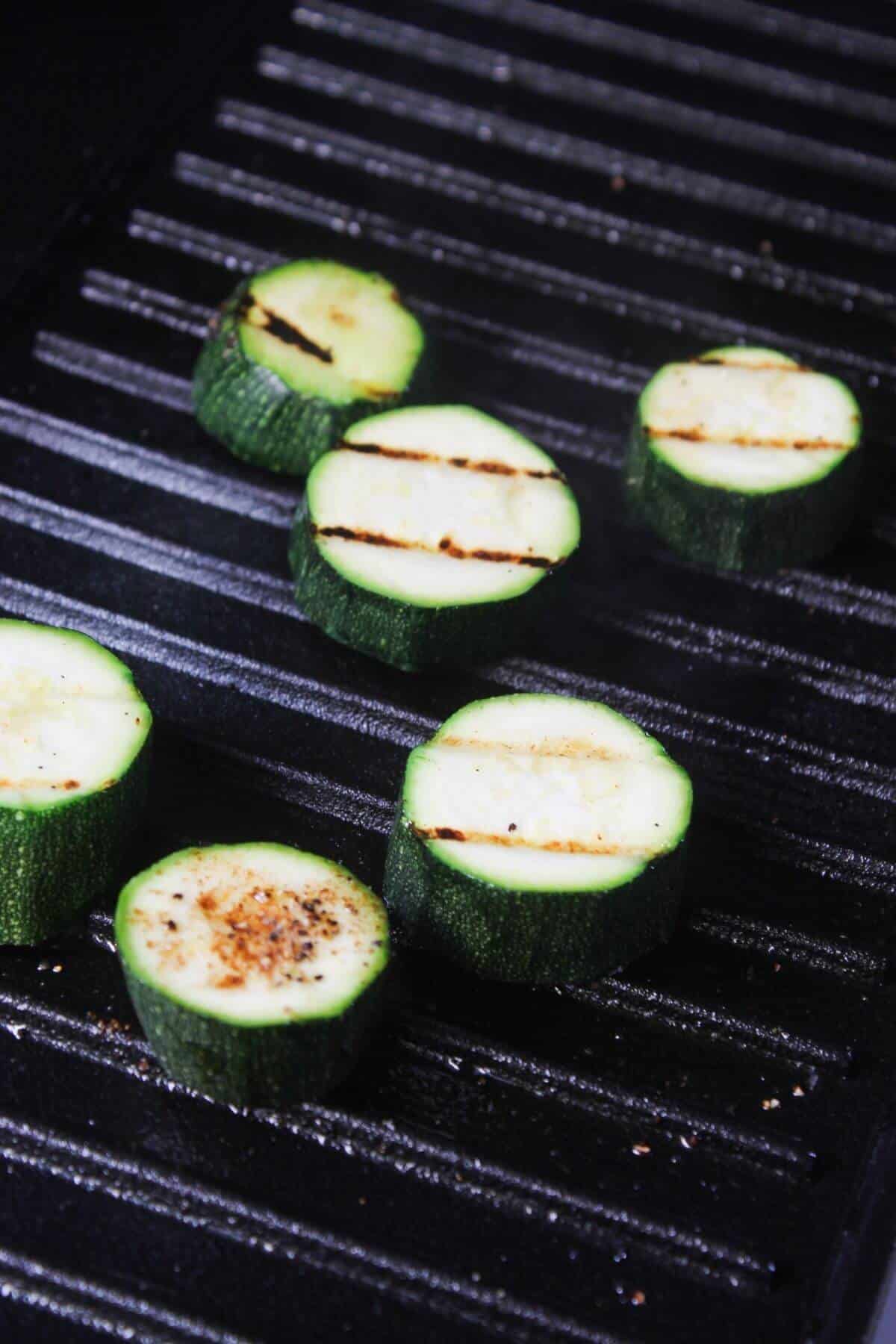 Sliced zucchini rounds cooking on a grill pan, showing char marks and light seasoning.