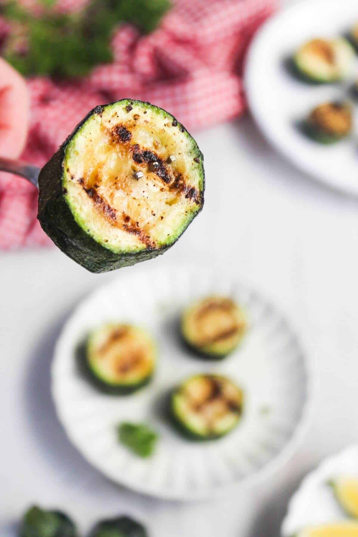 A close-up of a grilled zucchini slice held above a plate with more grilled zucchini pieces on a white surface.