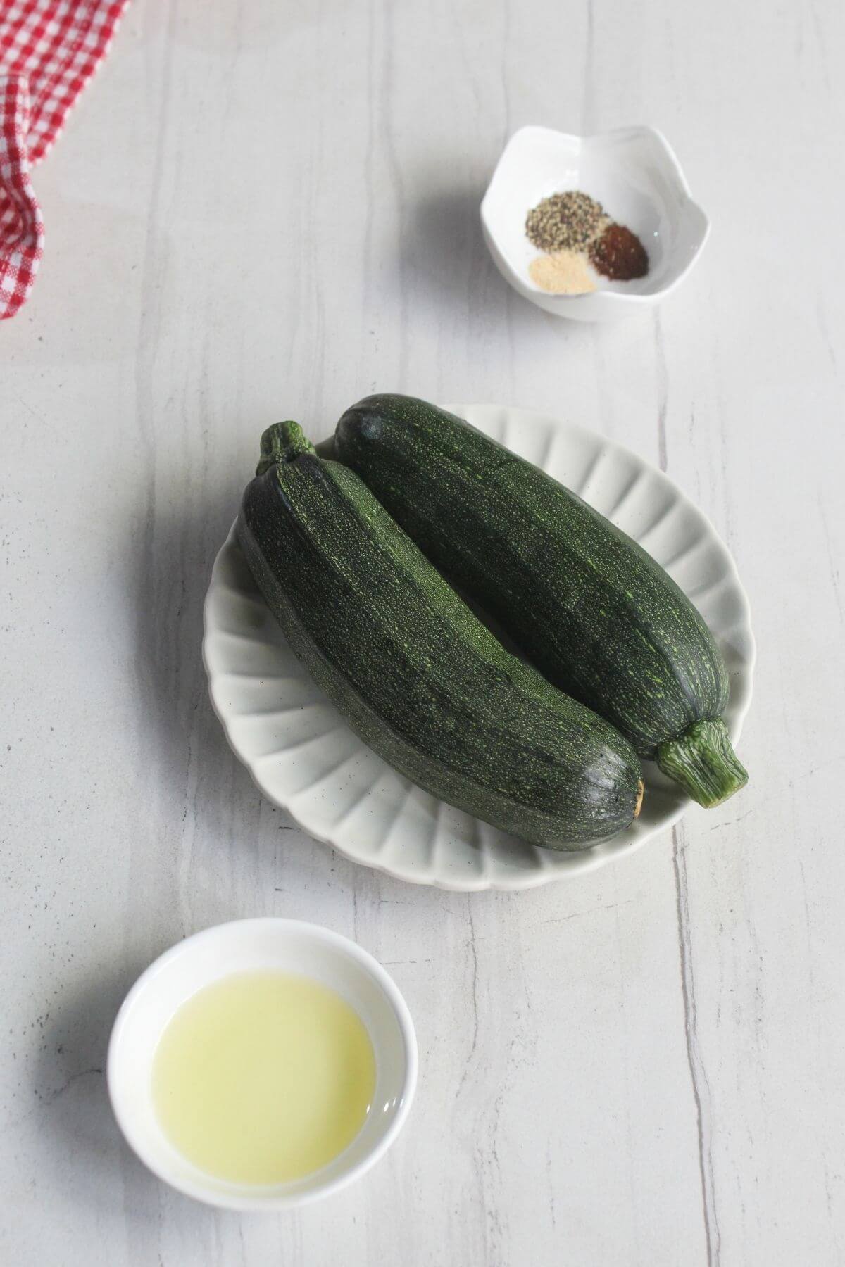 Two whole zucchinis on a white plate, with a small bowl of oil and a small bowl of assorted spices on a light-colored surface.