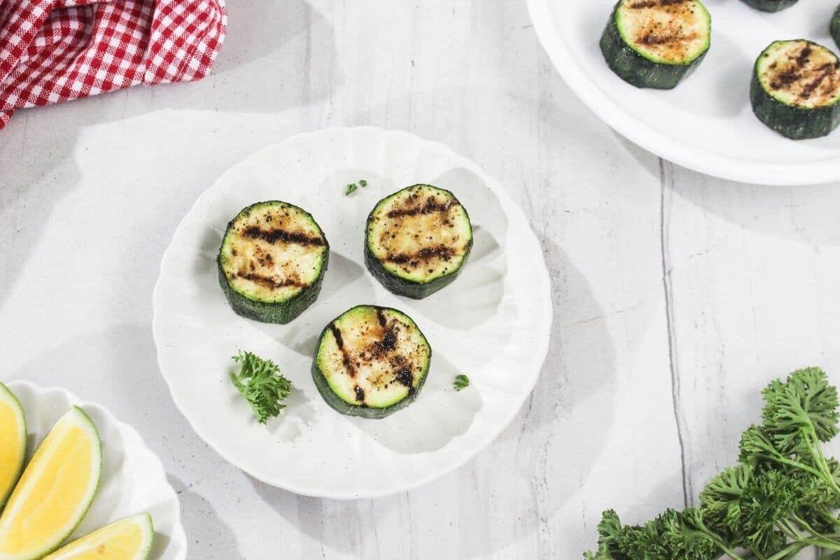 Three grilled zucchini slices on a white plate, garnished with parsley, with lemon wedges and a red checkered cloth nearby on a white surface.