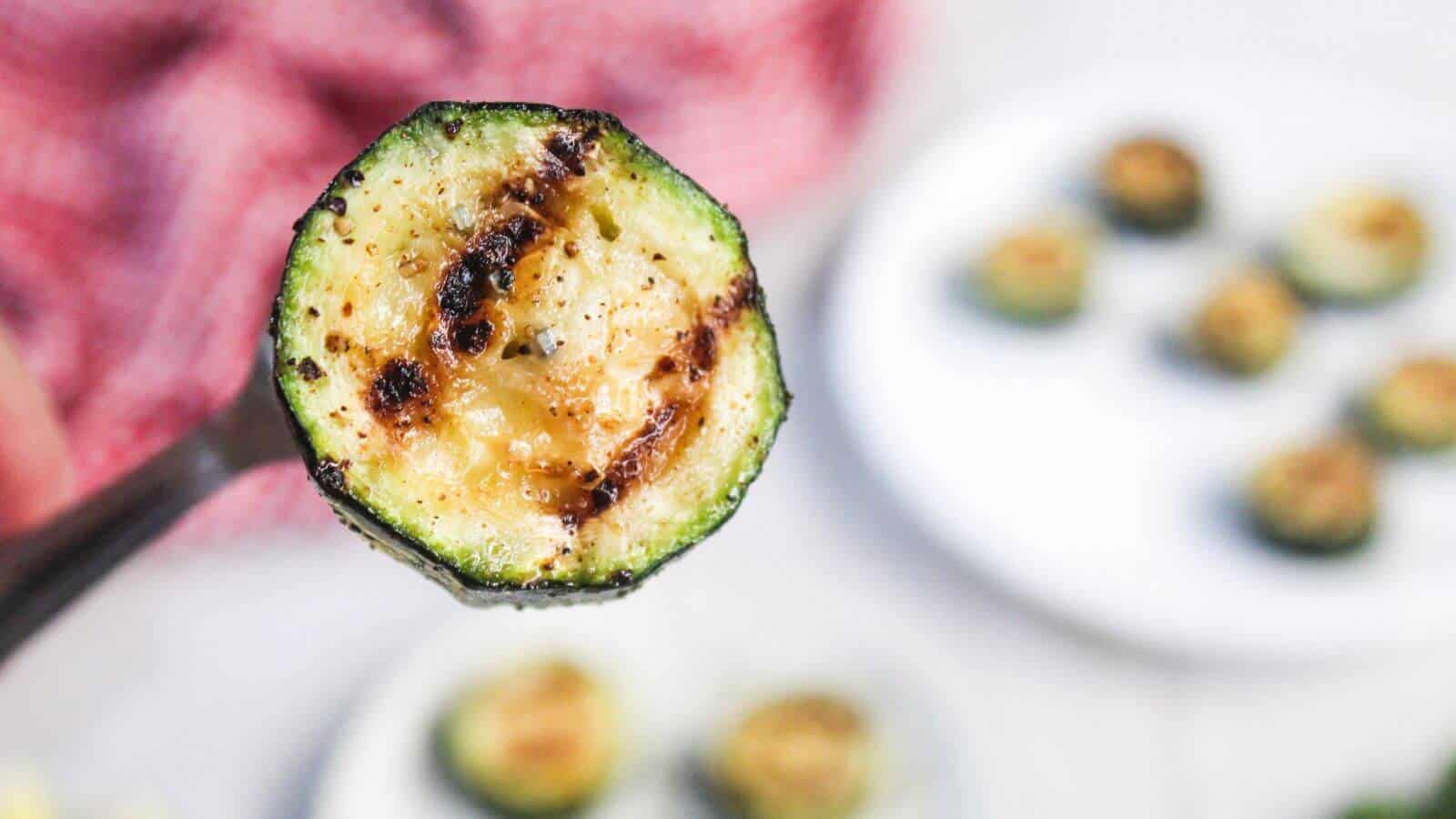 A close-up of a grilled zucchini slice on a fork, with more grilled zucchini pieces on a plate in the blurred background.