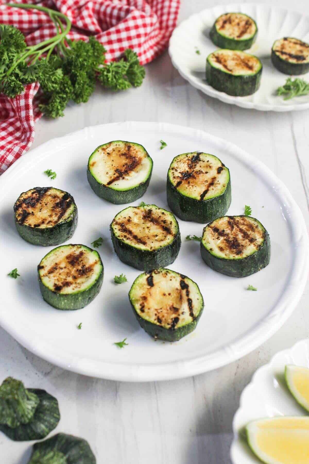 Grilled zucchini slices arranged on a white plate, garnished with chopped herbs, with a red checkered cloth and fresh parsley in the background.