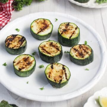 A white plate with seven grilled zucchini slices, each showing grill marks, garnished with small sprinkles of herbs.
