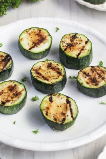 A white plate with seven grilled zucchini slices, each showing grill marks, garnished with small sprinkles of herbs.
