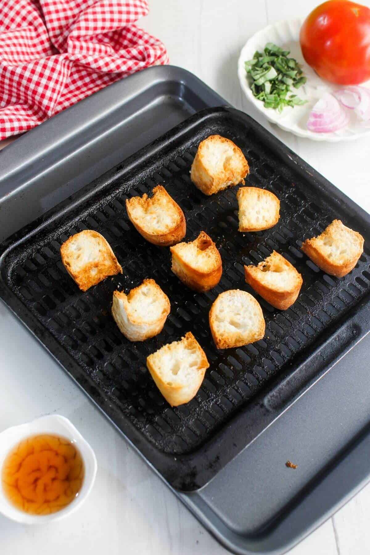 Sliced baguette pieces arranged on a grill pan, with a red checkered cloth, a tomato, herbs, shallot, and a small bowl of oil nearby on a white surface.