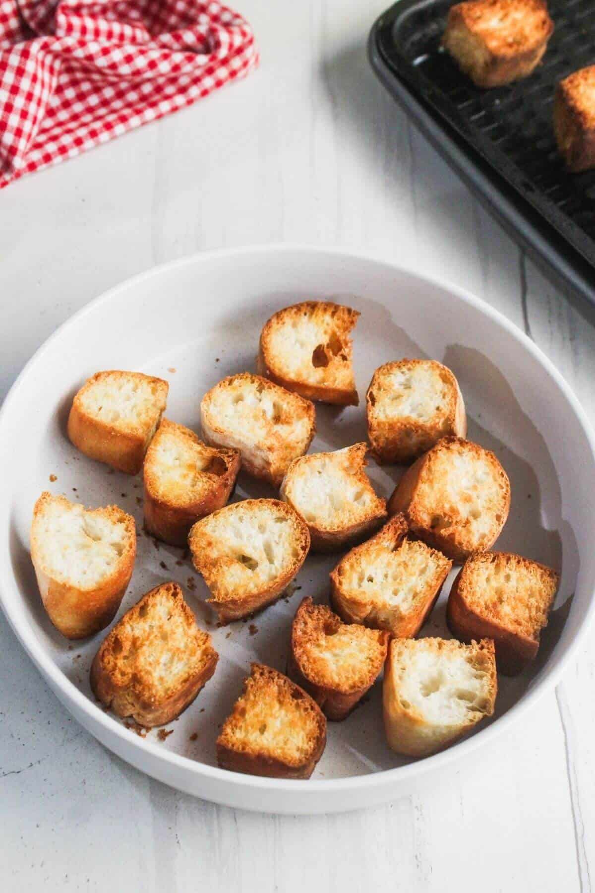 A white bowl filled with sliced, toasted pieces of baguette sits on a white surface; more toasted bread pieces are on a black tray in the background.