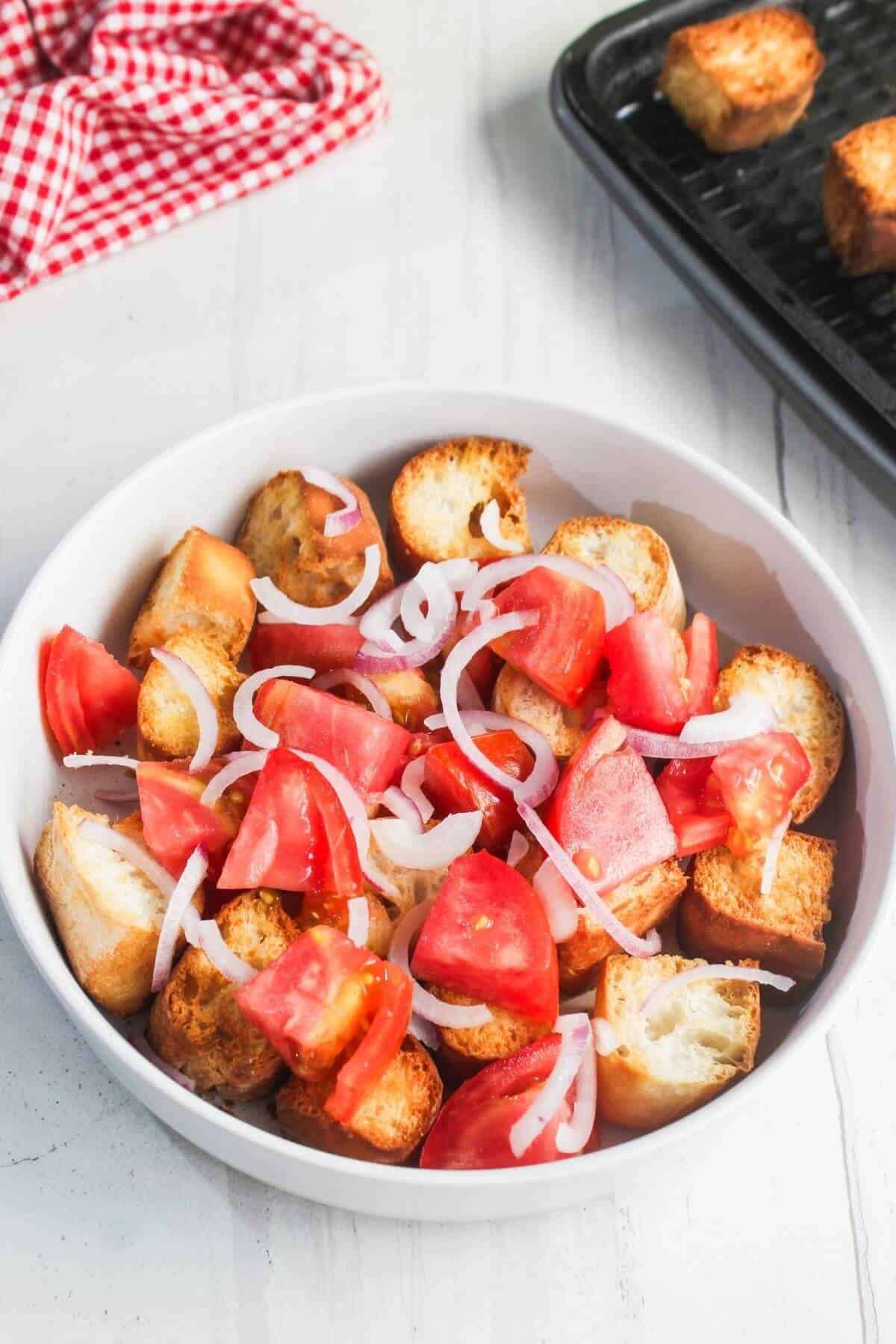 A white bowl filled with toasted bread cubes, sliced tomatoes, and thinly sliced onions on a light surface.