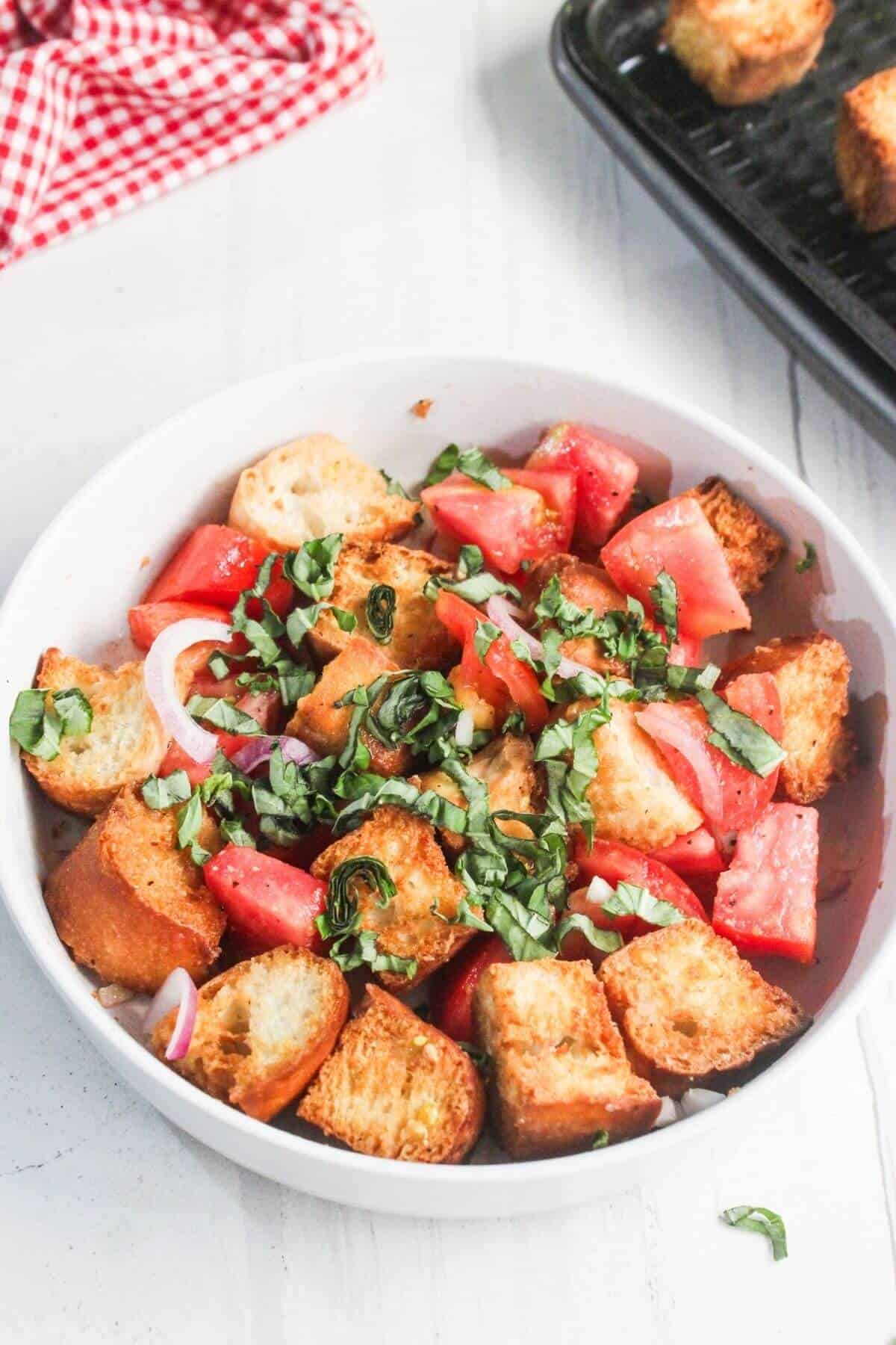 A white bowl filled with panzanella salad featuring chopped tomatoes, toasted bread cubes, sliced red onions, and fresh basil.