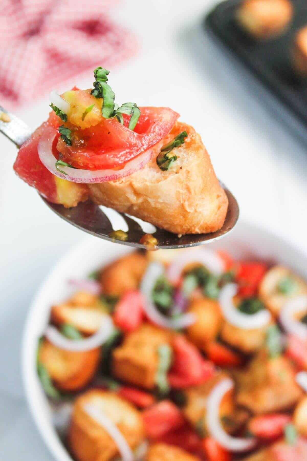 A spoon holds a piece of toasted bread topped with diced tomato, red onion, and chopped basil, with more similar pieces in a bowl below.