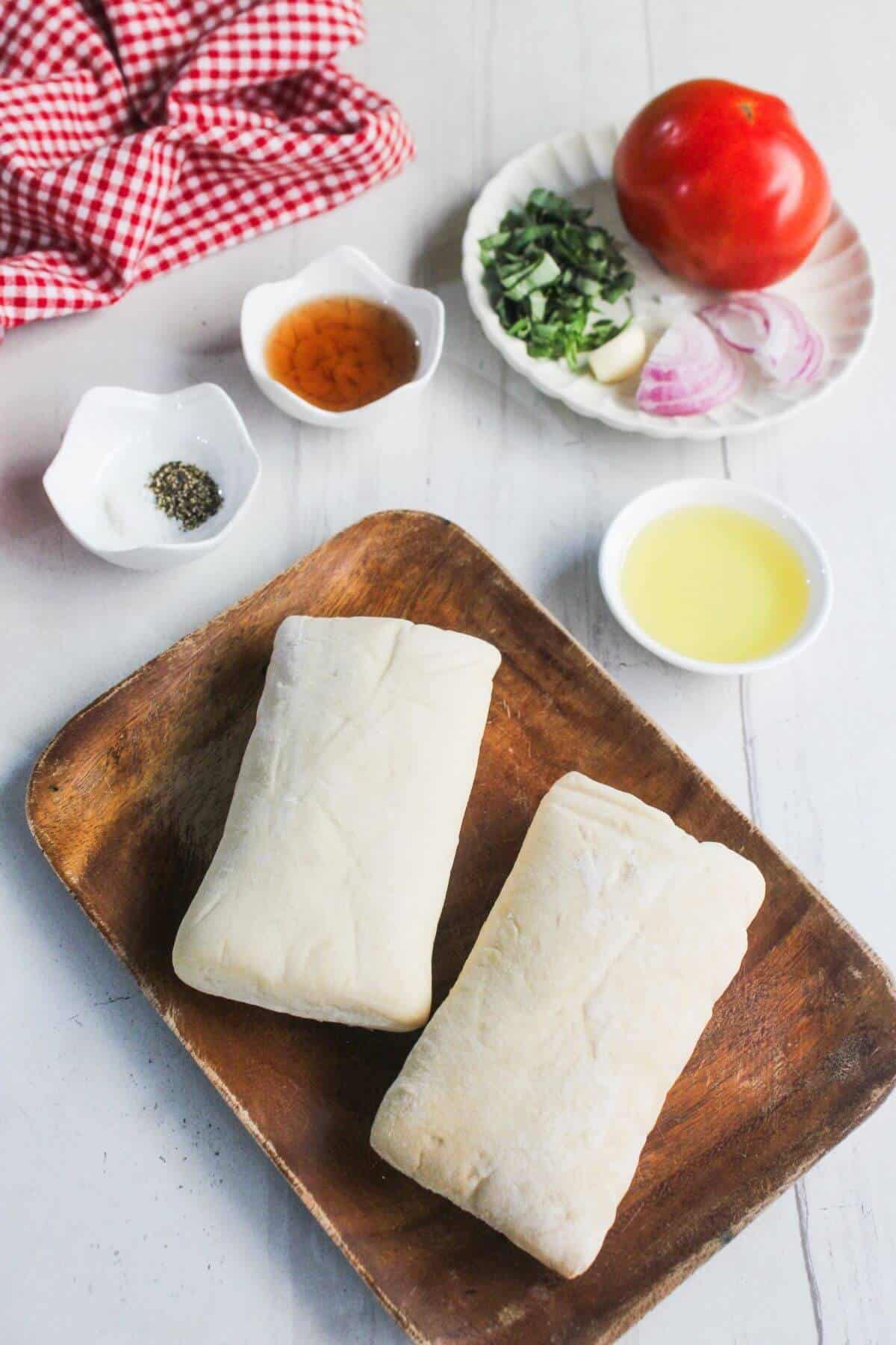 Two pieces of raw dough on a wooden plate, with small bowls of oil, vinegar, black pepper, sliced tomato, basil, garlic, and onion on a white table.