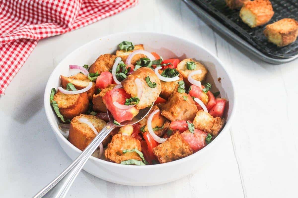 A white bowl filled with panzanella salad, featuring toasted bread cubes, tomato chunks, sliced onions, and basil, with serving utensils inside.