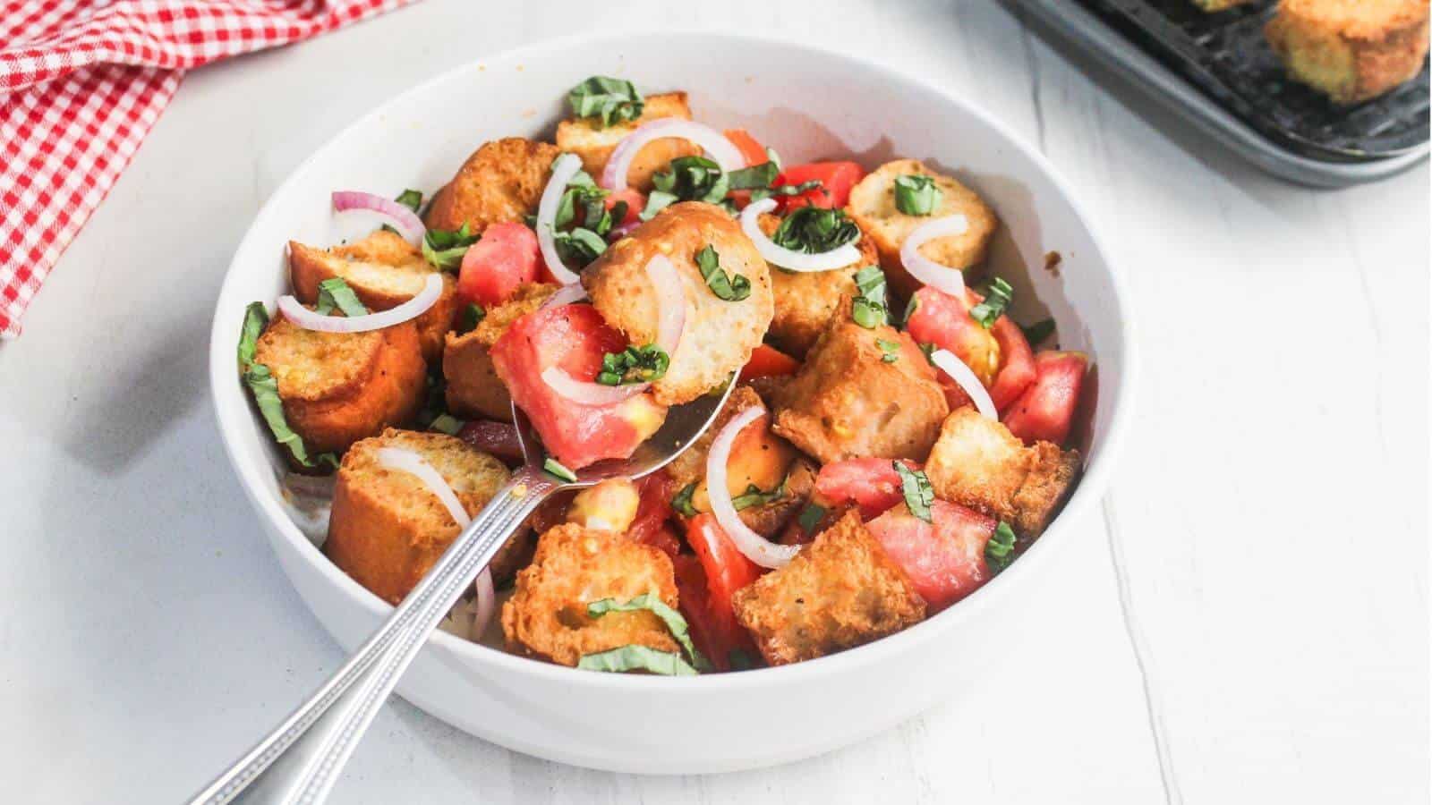 A white bowl filled with panzanella salad containing bread cubes, tomato chunks, sliced red onion, and fresh basil, with a spoon resting inside the bowl.