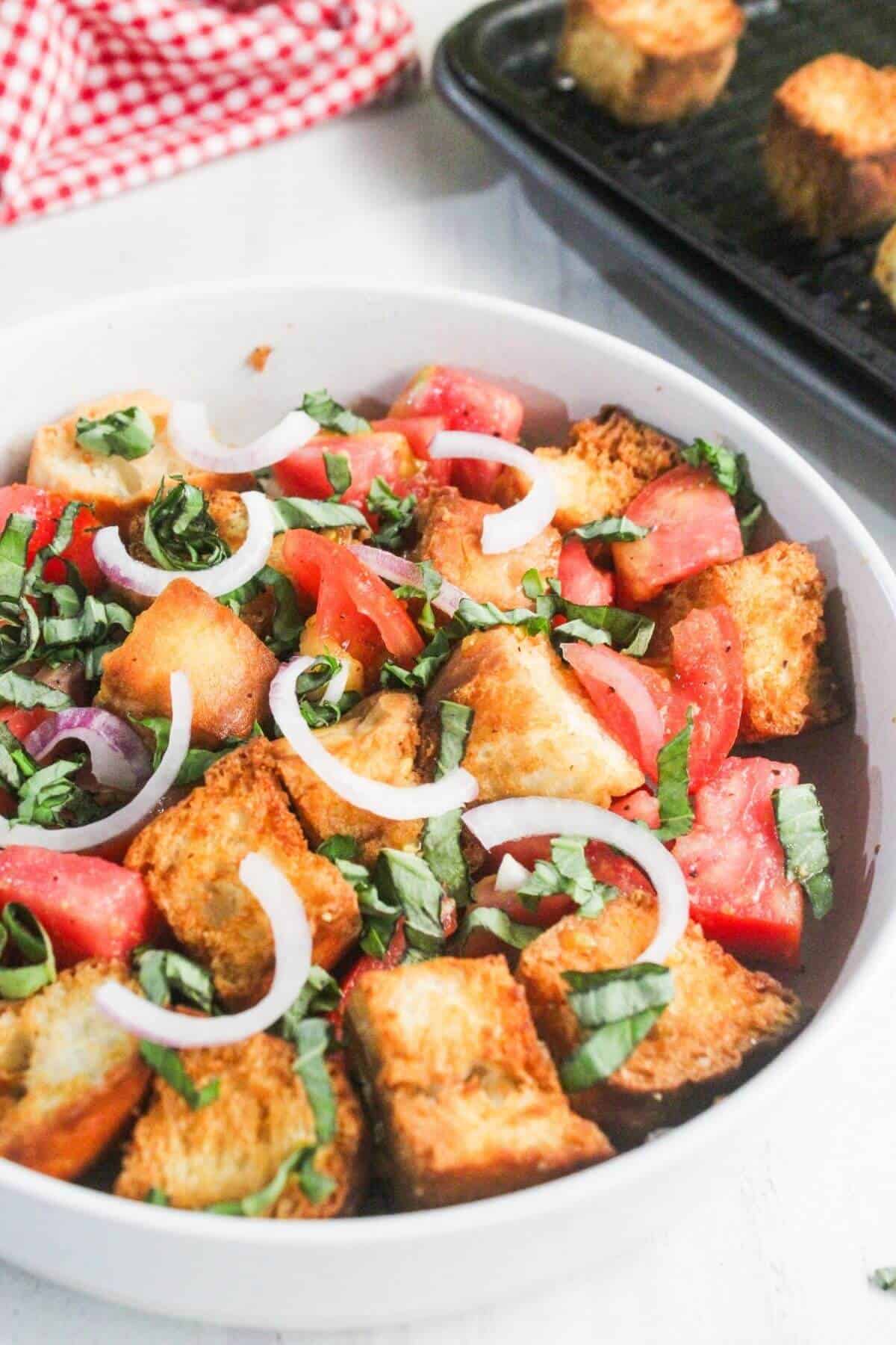 A bowl of panzanella salad with chunks of bread, tomato, onion slices, and fresh basil, next to a tray of toasted bread pieces.