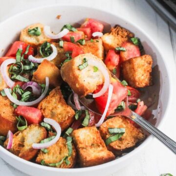 A bowl of panzanella salad with toasted bread cubes, chopped tomatoes, sliced red onions, and fresh basil, with a spoon inside the bowl.