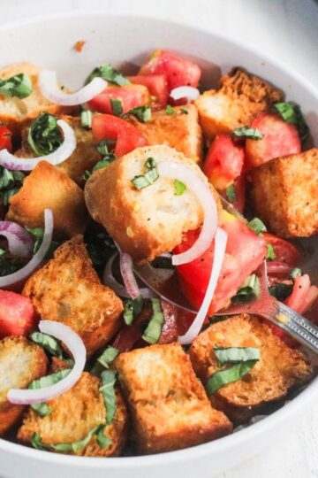 A bowl of panzanella salad with toasted bread cubes, chopped tomatoes, sliced red onions, and fresh basil, with a spoon inside the bowl.