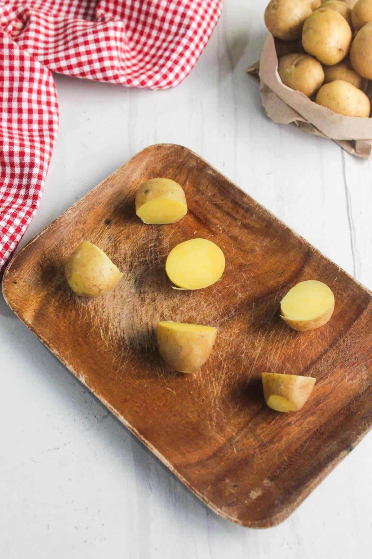 A wooden tray with five cut pieces of yellow potatoes on a white surface, a red and white checkered cloth, and a small basket of whole potatoes in the background.