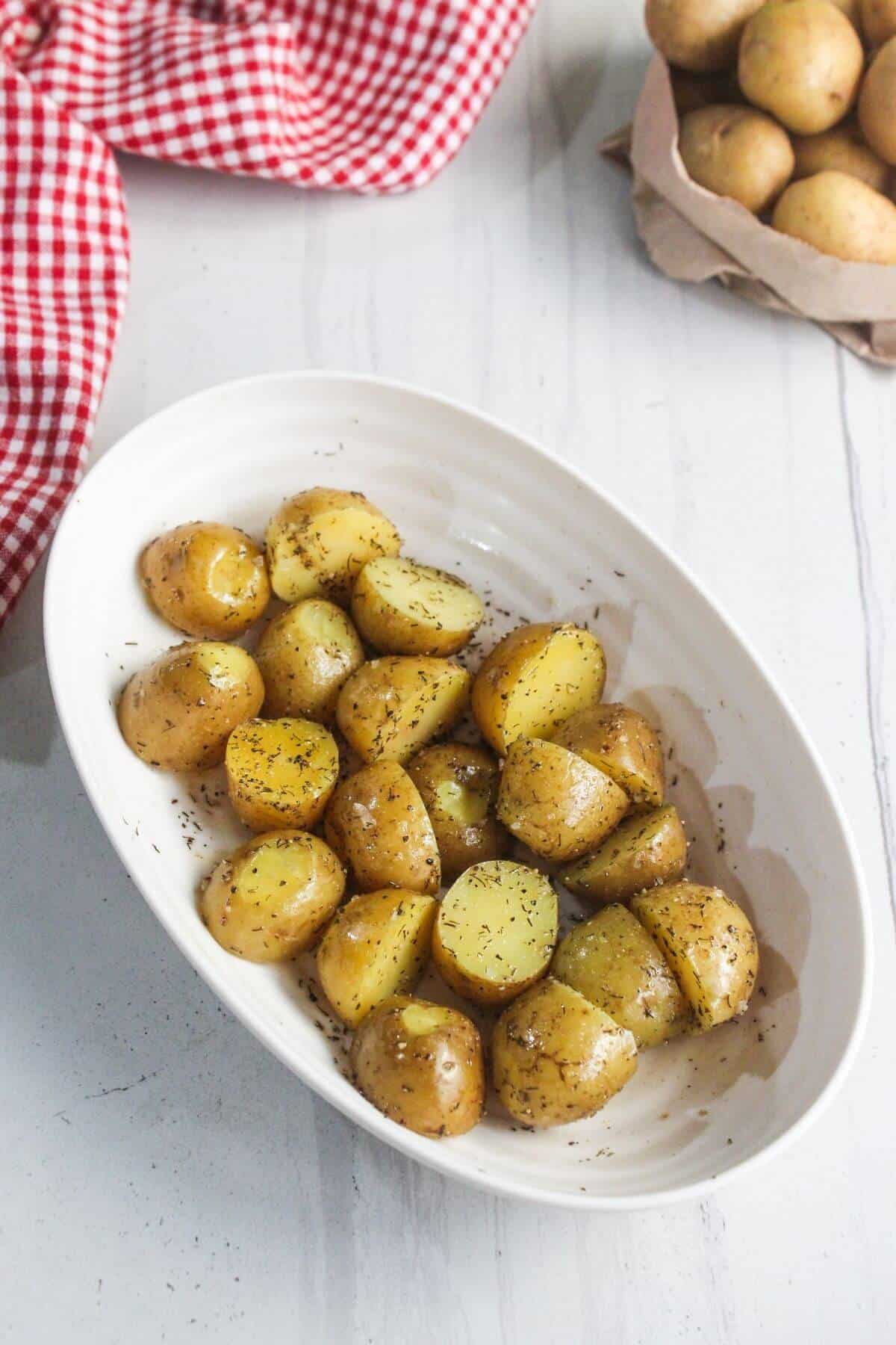 A white oval dish filled with seasoned, roasted baby potatoes sits on a white surface near a red checkered cloth and a bag of raw potatoes.