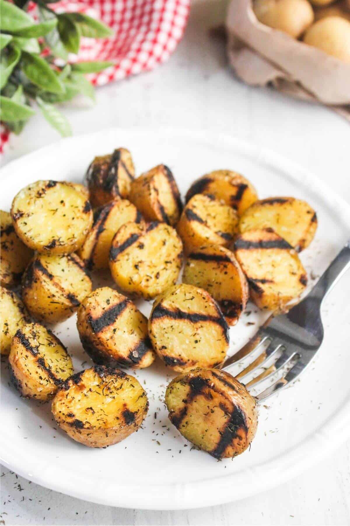 Grilled potato halves with grill marks, seasoned with herbs, served on a white plate with a fork.