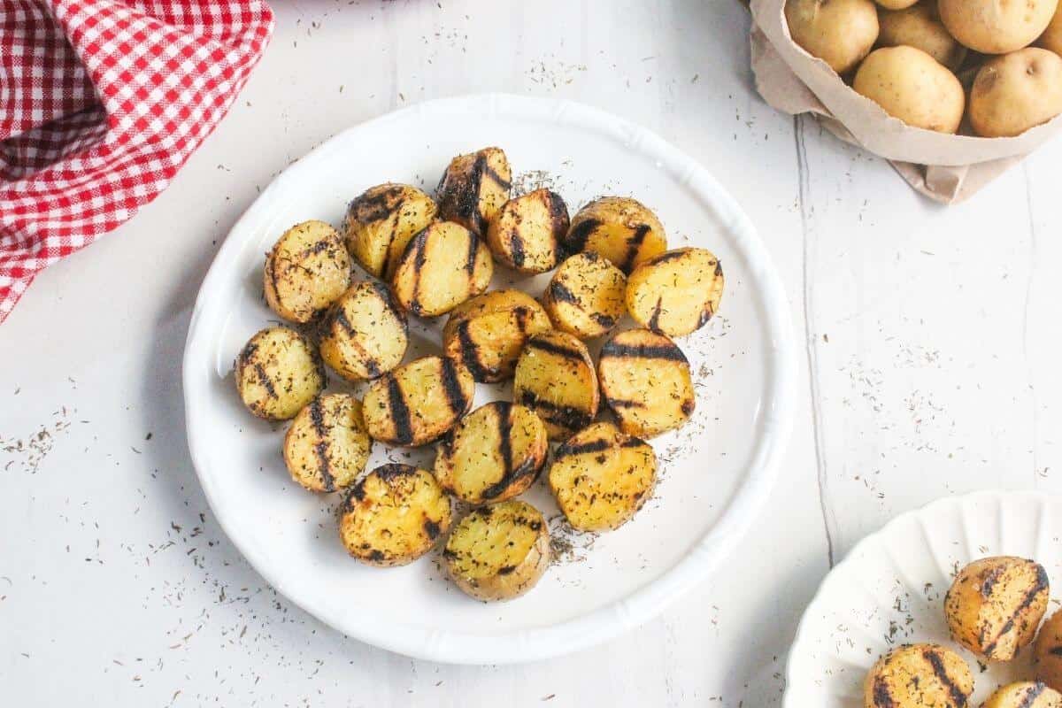 A white plate holds grilled potato halves with char marks, surrounded by fresh potatoes, a red checked cloth, and a white plate on a white surface.