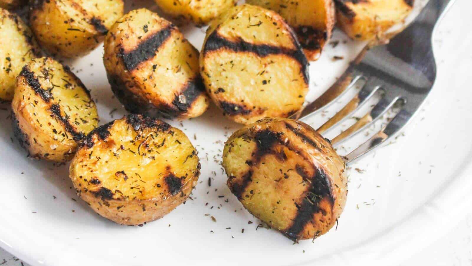 Grilled potato slices with seasoning on a white plate, with a fork resting beside them.