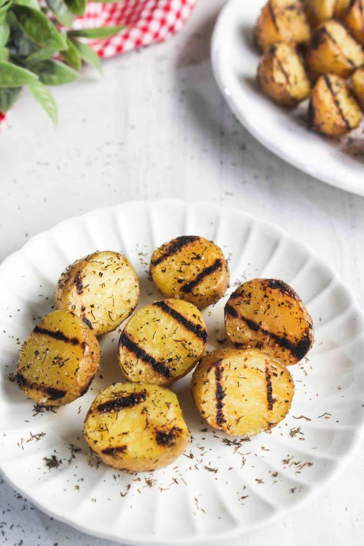 A white plate with six grilled potato halves seasoned with herbs, showing char marks, on a white surface with more potatoes in the background.